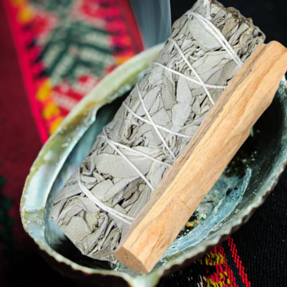 Bundles of sage tied with twine on a metal tray with a wooden tool, against a colorful embroidered fabric background.