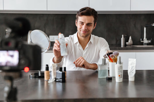 A man in a kitchen holding a bottle of skincare product, with a camera and makeup products on the counter.