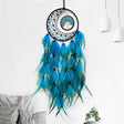 Blue and green dreamcatcher hanging on a white wall with a gray pillow and plant in the background.