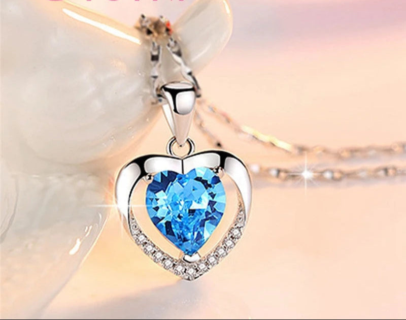 Heart-shaped pendant with blue gemstone and clear stones on a soft background, featuring the brand 'Giemi'.