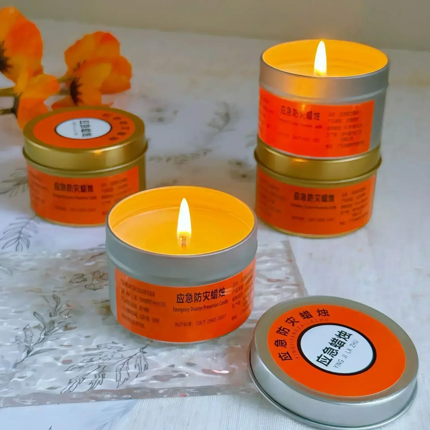 Candle tins with orange labels and silver lids on a white surface with flowers.