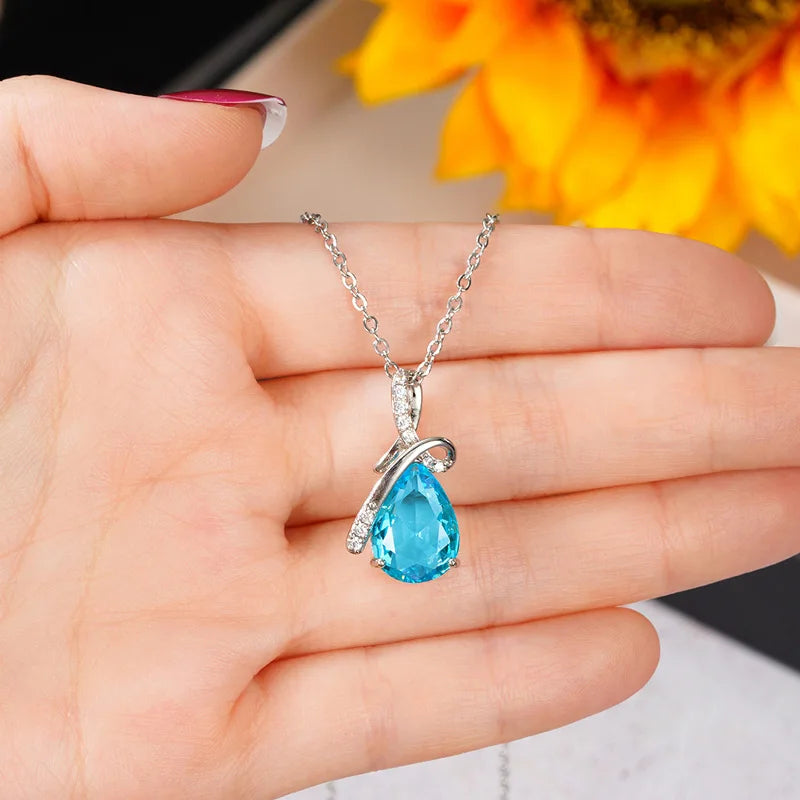 Blue teardrop pendant on a silver chain held in a hand with a blurred sunflower background