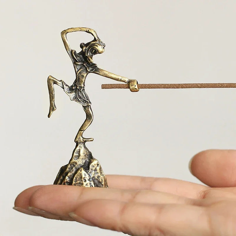 Small bronze sculpture of a figure on a stick held in a hand against a plain background