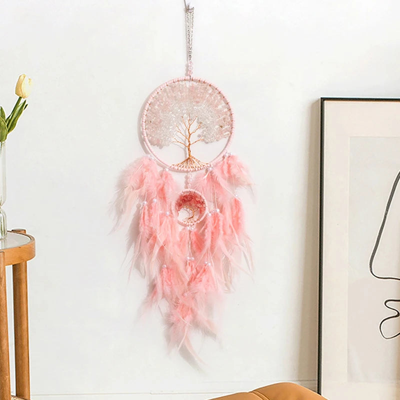 Pink dreamcatcher with feathers hanging on a white wall next to a framed picture and small table.