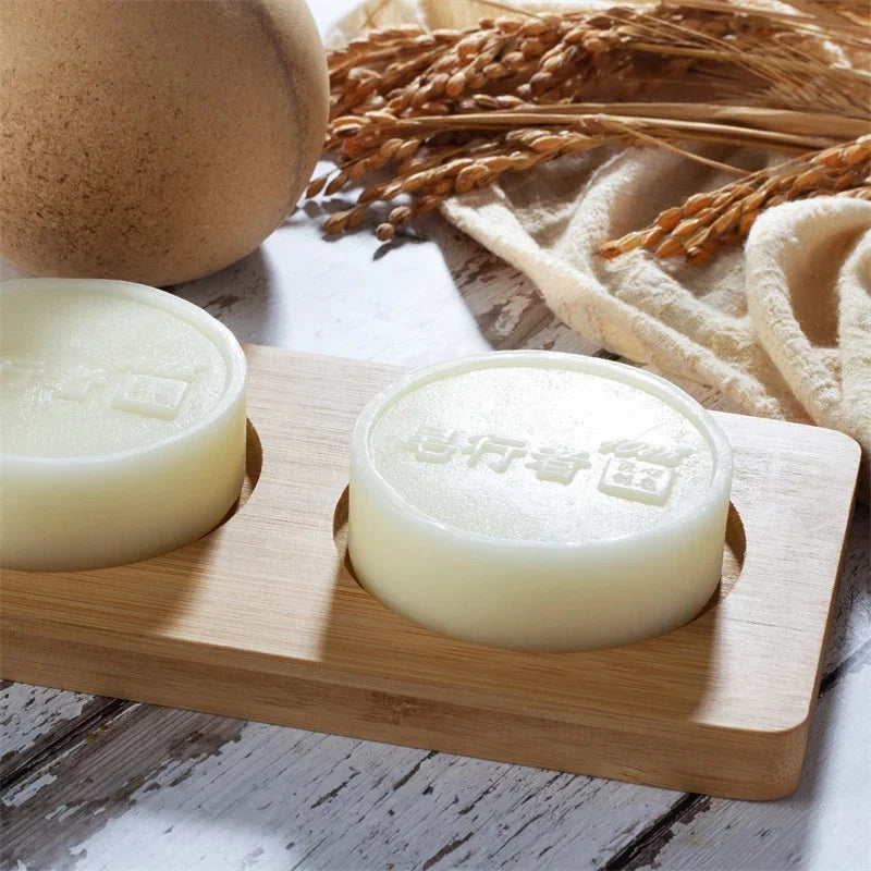 Two soaps on a wooden soap dish with a rustic background featuring a loaf of bread and wheat stalks.