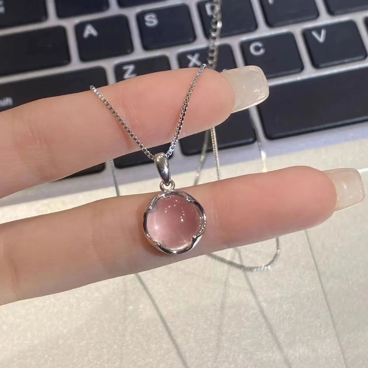 Silver necklace with a pink pendant held by a hand in front of a laptop keyboard.