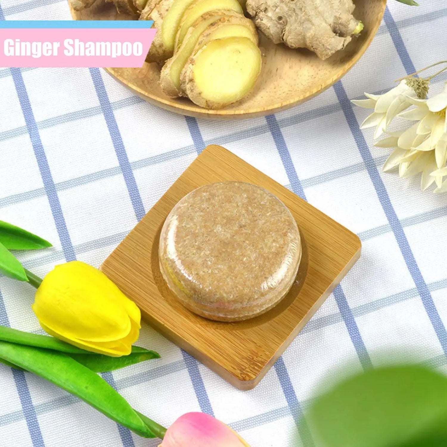 Ginger shampoo bar on a wooden coaster with ginger slices and flowers on a checkered tablecloth.