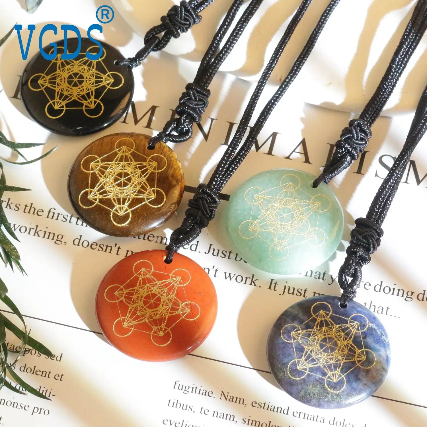 Five stone pendants with geometric designs on black cords, branded 'VCGDS', on a magazine background.