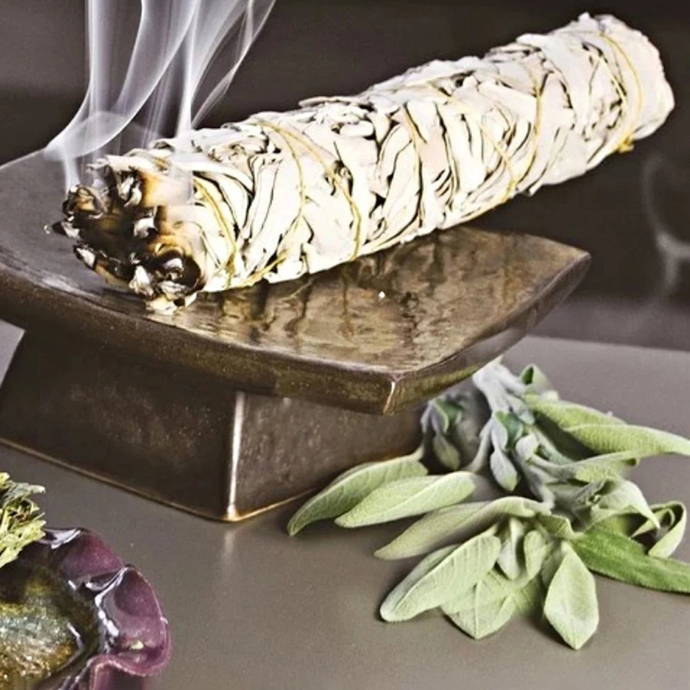 Sage smudge stick on a stone holder with smoke and sage leaves on a dark surface