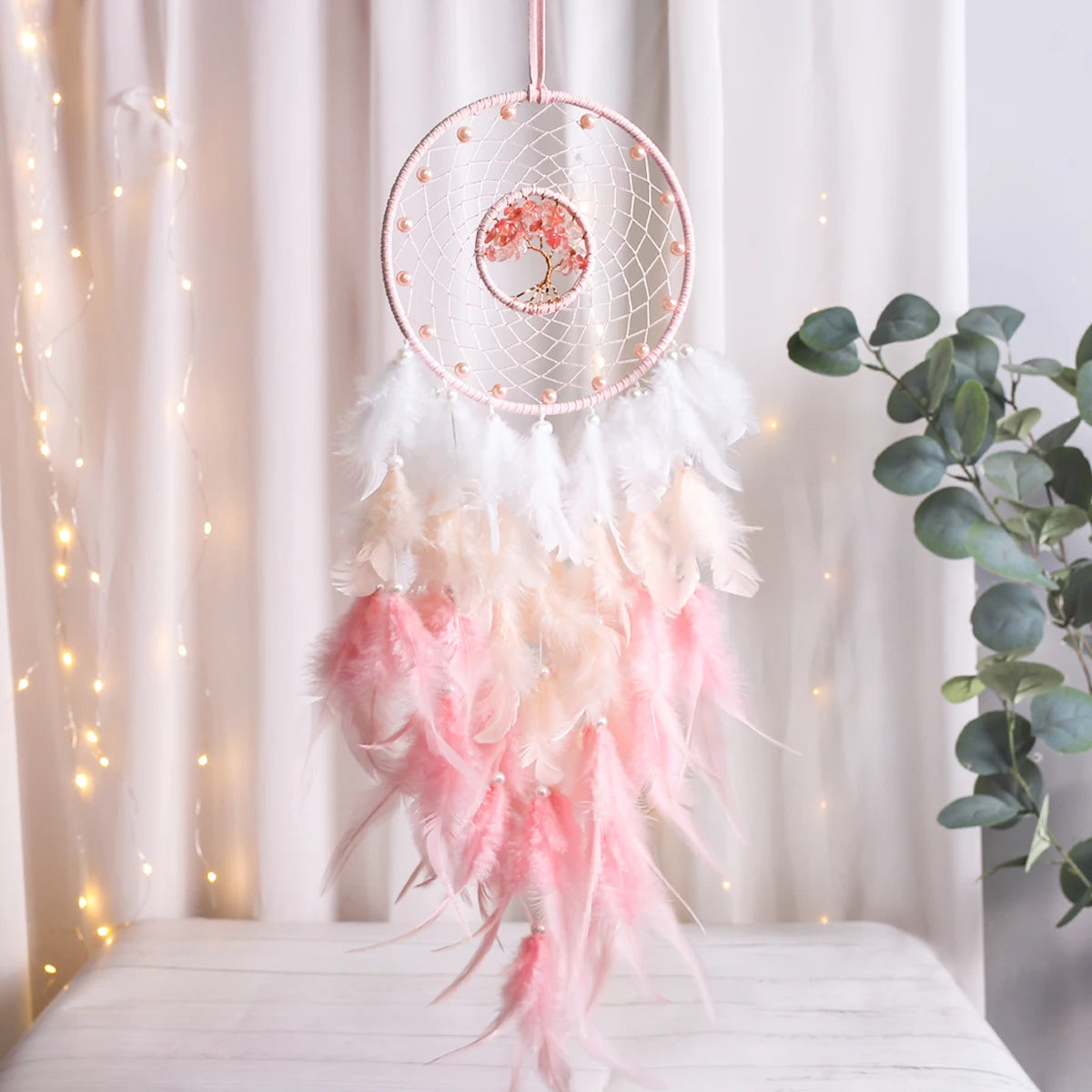 Pink dreamcatcher with feathers on a white surface with fairy lights and a plant in the background