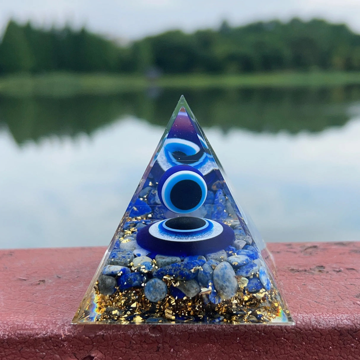 Pyramid-shaped crystal with blue and gold elements on a red surface with a blurred natural background