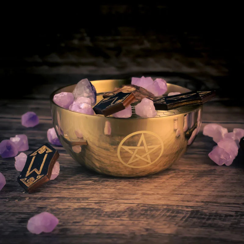 Gold bowl with pentagram design on a wooden surface surrounded by crystals and wands.