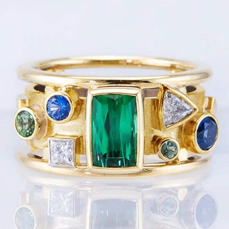 Gold ring with green emerald, blue sapphire, and white diamond gemstones on a reflective surface.