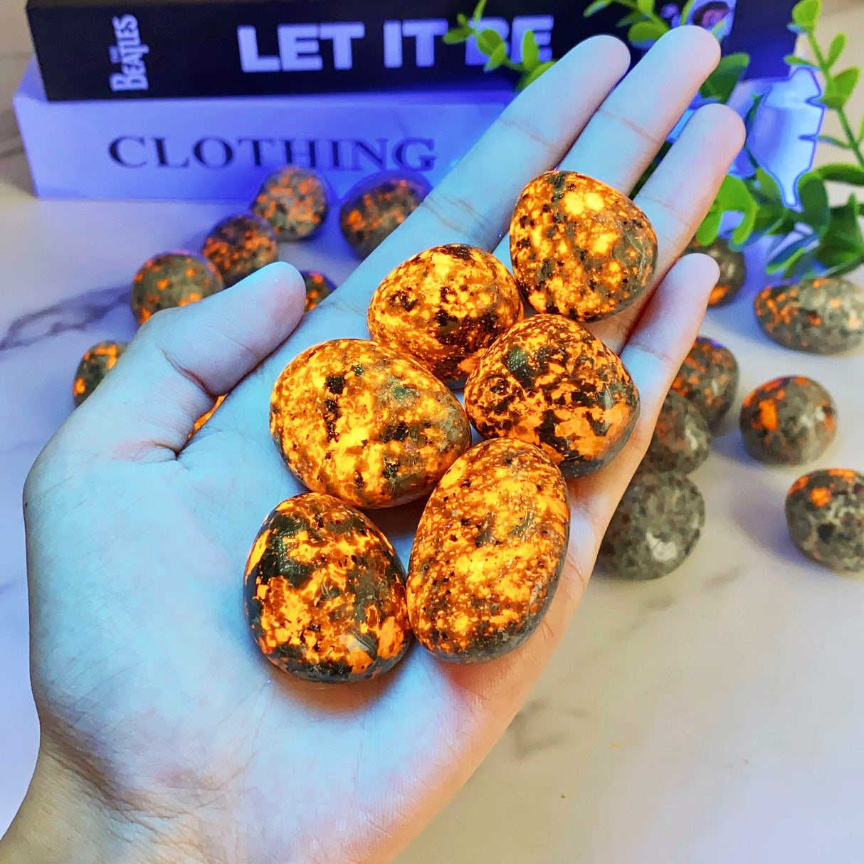 Hand holding a small pile of metallic orange and black objects with 'Let It Be' clothing box in the background.