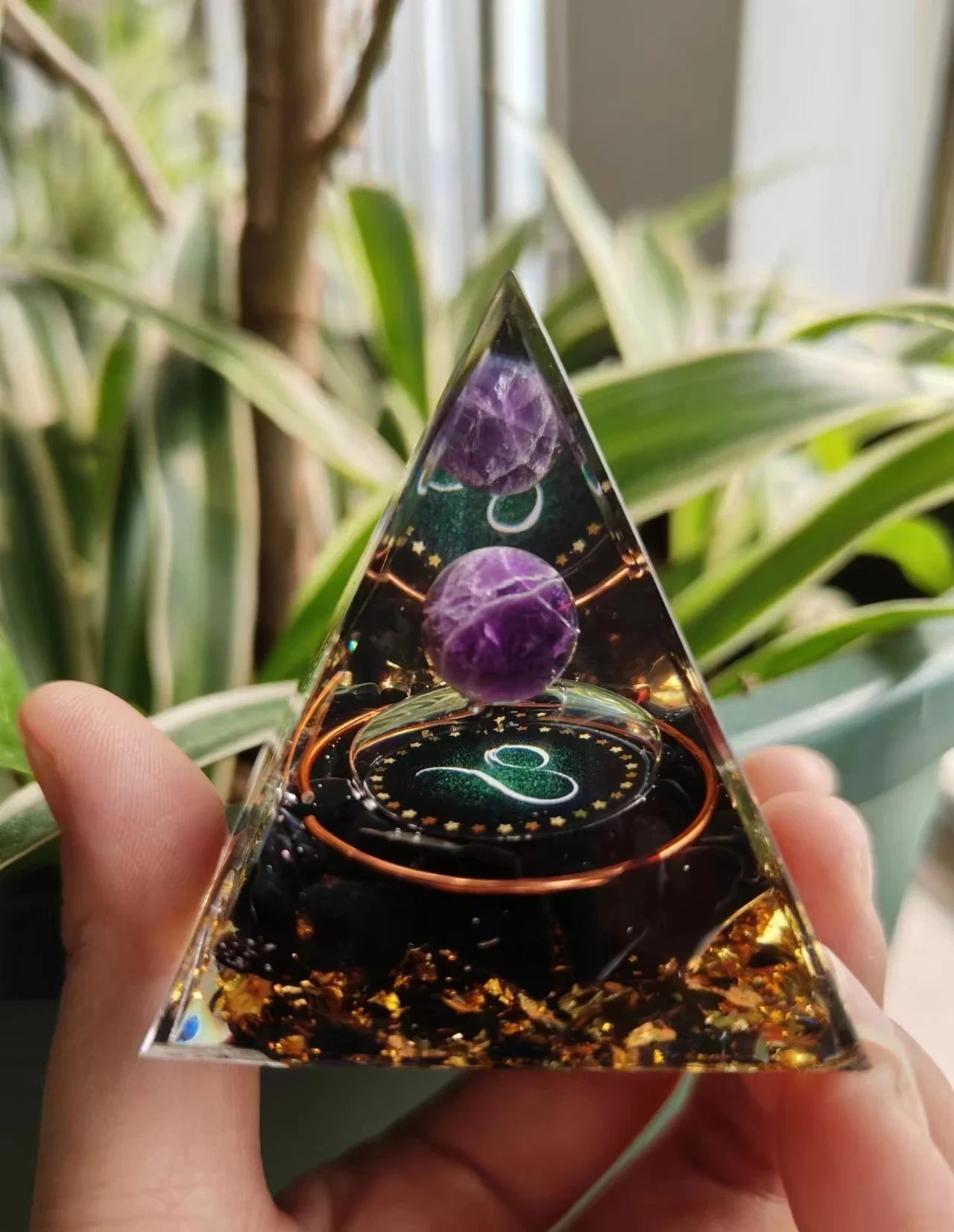 Triangular crystal pyramid with purple sphere held by a hand, against a blurred green plant background