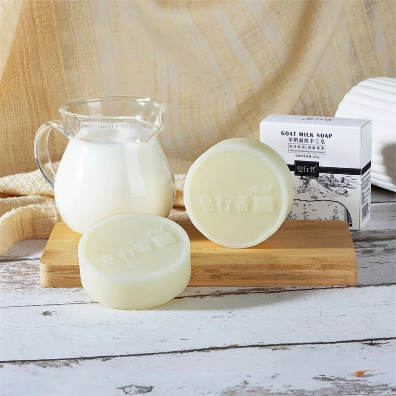 Three bars of goat milk soap on a wooden block with a container of milk and a box labeled 'GOAT MILK SOAP' in the background.