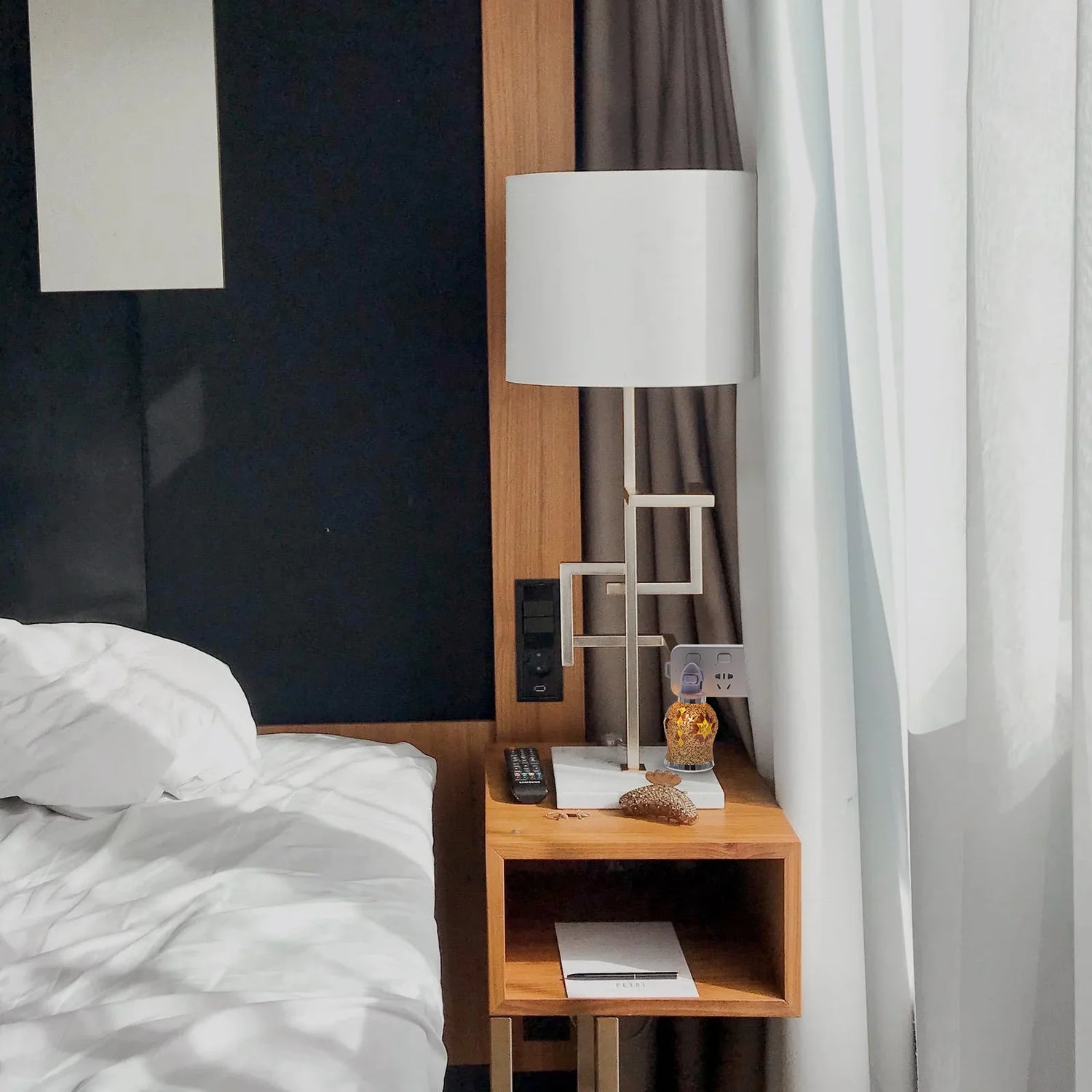 Wooden nightstand with lamp and remote control next to a bed in a room with curtains.