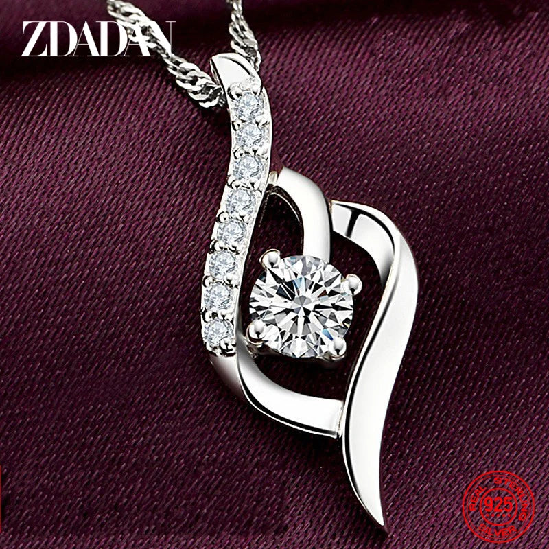 Silver heart-shaped pendant with clear gemstone on a maroon fabric background, featuring the brand 'ZDADAN'.
