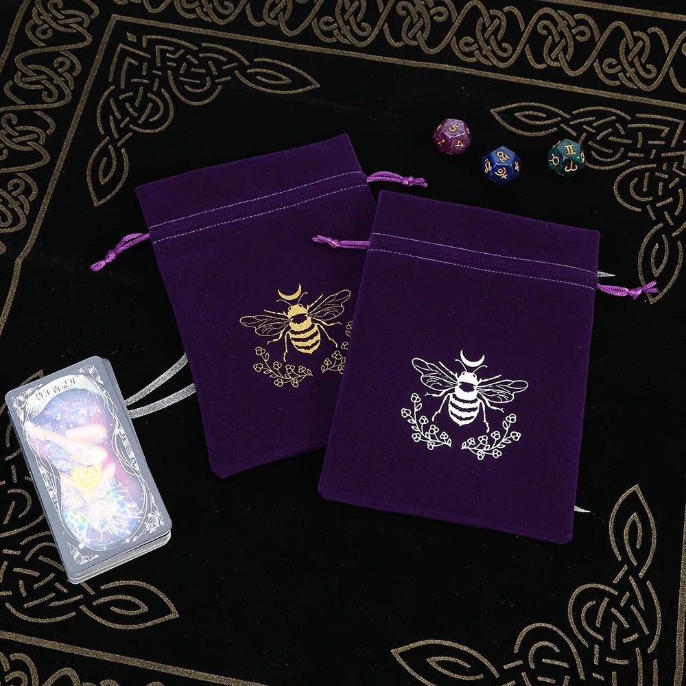 Two purple velvet bags with bee designs on a black background with tarot cards and dice.