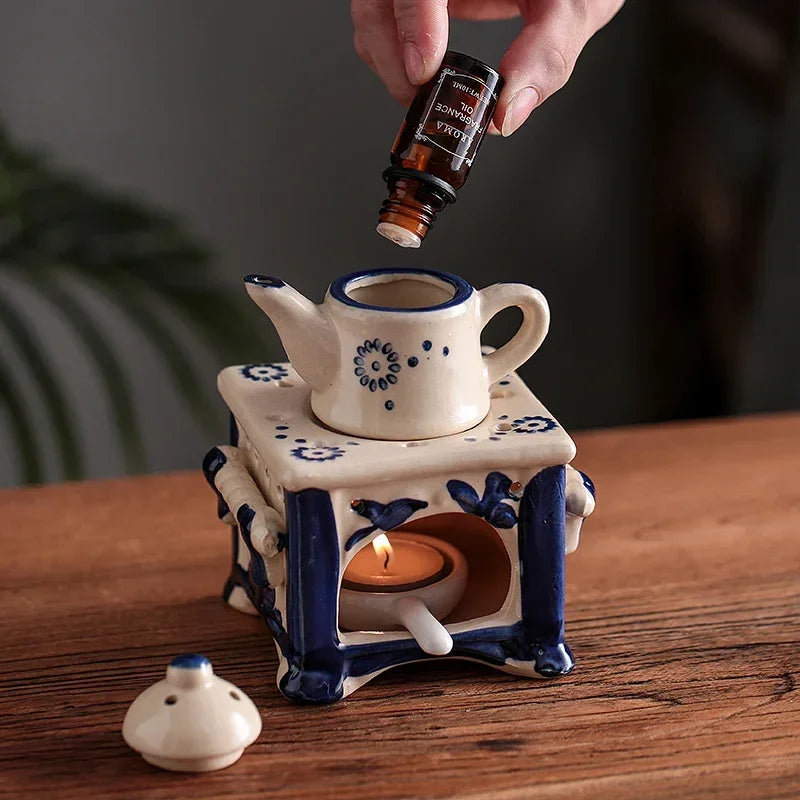 Ceramic oil burner with a lit candle and a hand holding a bottle of essential oil above it on a wooden surface.