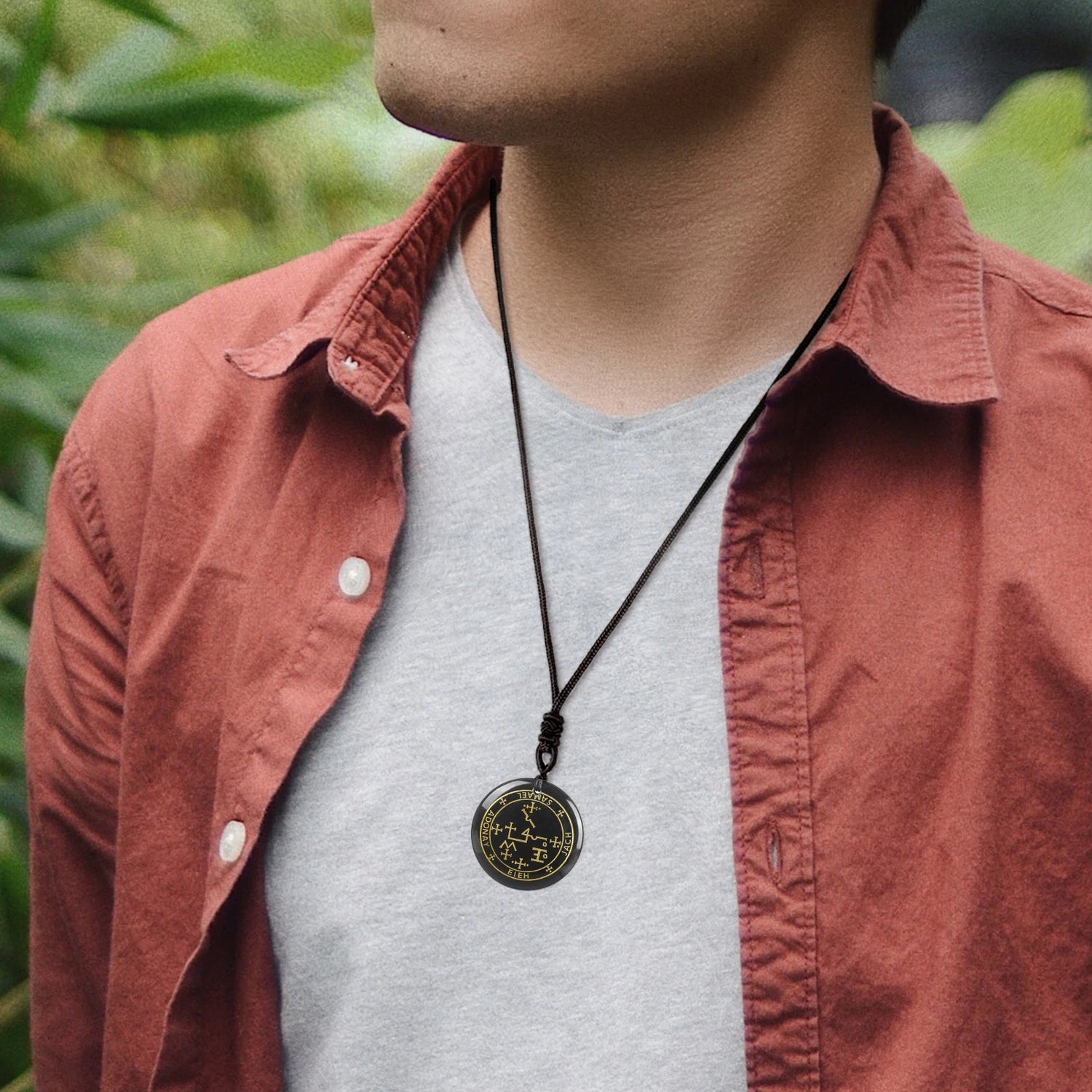 Person wearing a necklace with a pendant, standing outdoors with greenery in the background