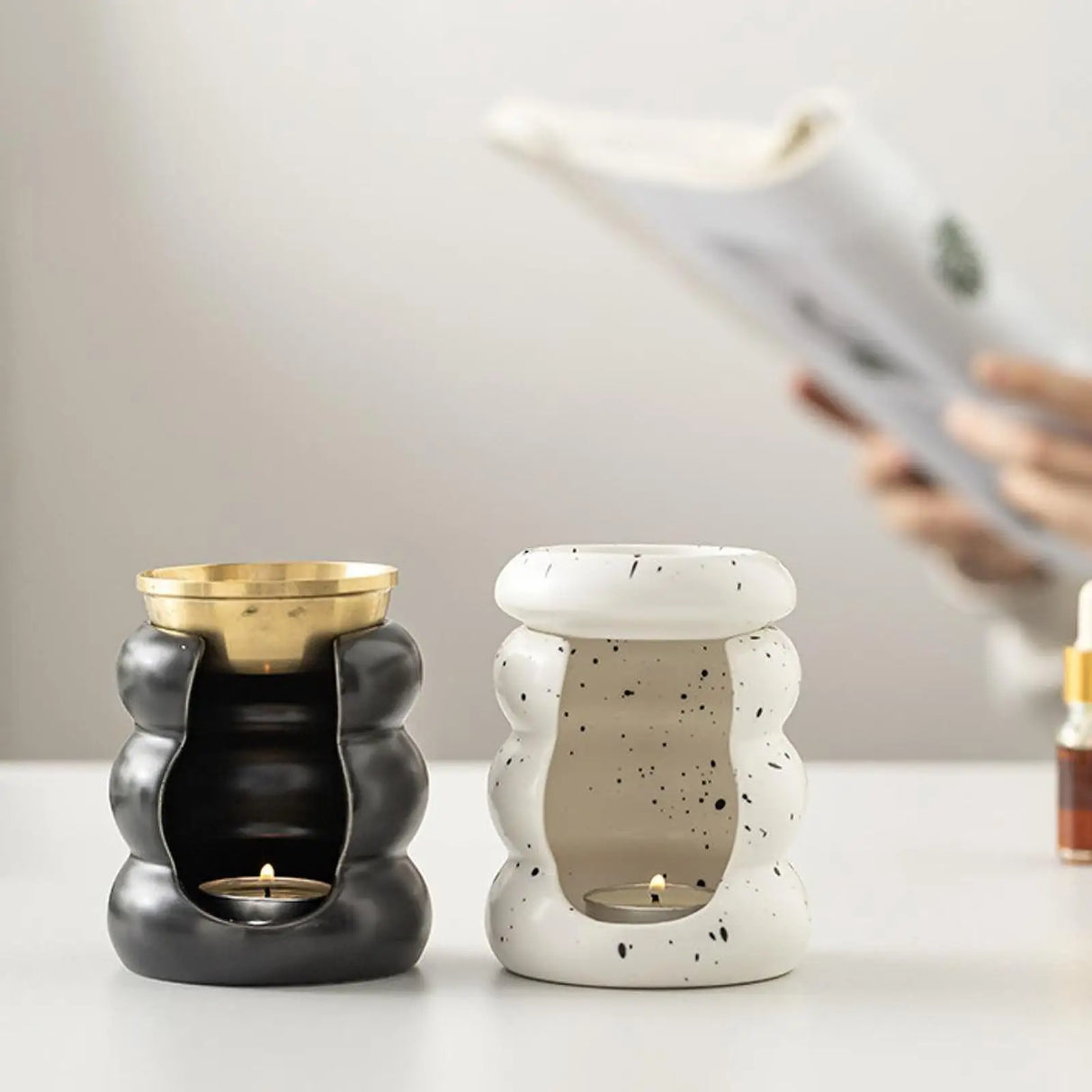 Two ceramic wax warmers, one black and gold, the other white with speckles, on a table.