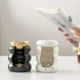 Two ceramic wax warmers, one black and gold, the other white with speckles, on a table.