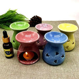 Set of colorful ceramic oil burners with a lit candle and essential oil bottle on a textured surface.