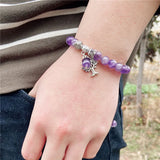 Purple beaded bracelet with a silver tree charm on a wrist, blurred outdoor background