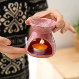 Pink ceramic oil burner with a lit candle inside, held by hands against a blurred background.