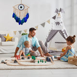 Man and two children playing with a toy train set in a room with a teepee and decorative wall hanging.