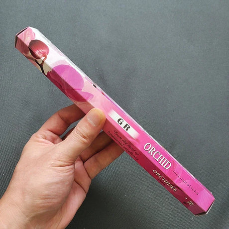 Hand holding a pink orchid incense stick box against a gray background