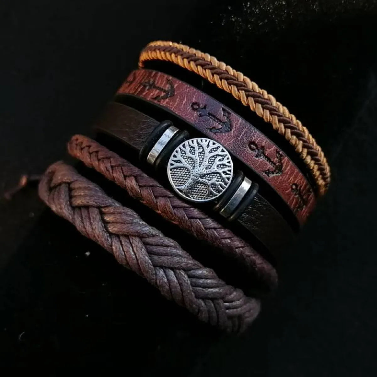 Stack of braided leather bracelets with a central silver tree-of-life charm on a dark background