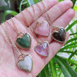 Heart-shaped gemstone pendants on gold chains held in a hand with a green plant background