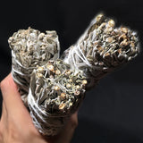 Three bundles of sage wrapped in white fabric held by a hand against a dark background