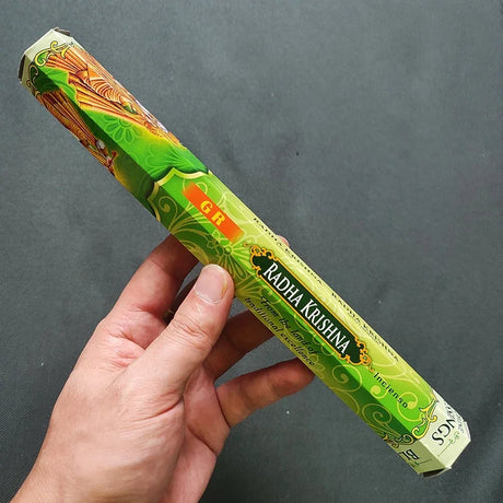 Hand holding a green 'Radha Krishna' incense stick packaging against a gray background