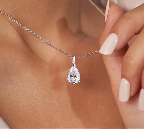 Teardrop-shaped diamond pendant on a chain, held by a hand with a blurred background.