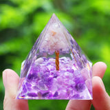 Pyramid-shaped crystal with purple and white stones held by a hand against a green blurred background