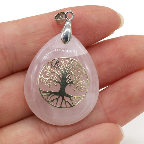 Pink teardrop-shaped pendant with gold tree design held in a hand against a white background