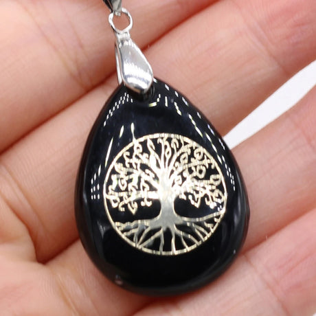 Black teardrop-shaped pendant with silver tree of life design held in a hand.