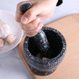 Person using a black speckled mortar and pestle on a wooden surface with garlic cloves nearby.