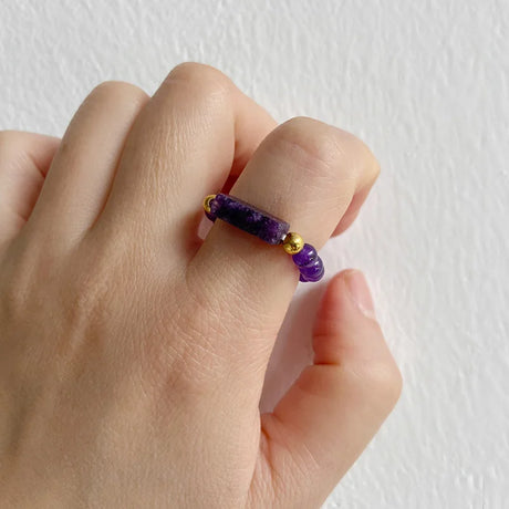 Hand wearing a purple beaded ring with gold accents on a white background