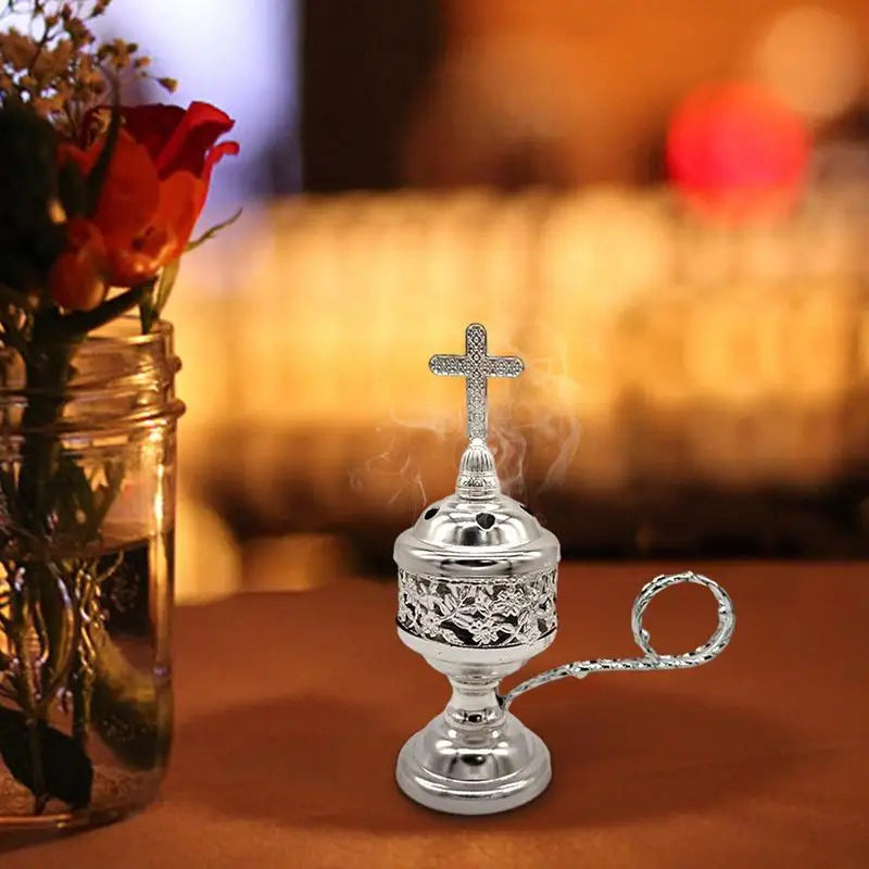 Silver incense burner with a cross on top, emitting smoke, next to a vase of red flowers.
