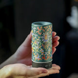 Hand holding a cylindrical ceramic container with intricate floral patterns against a dark background