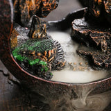 Decorative incense burner with rocks and greenery, emitting smoke.