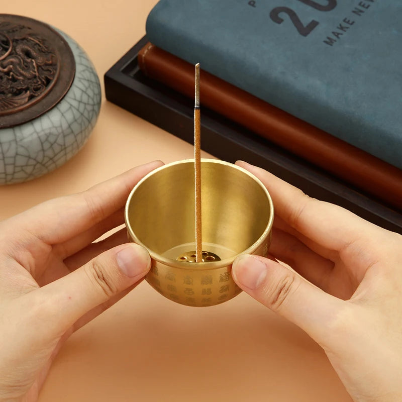 Gold incense burner with a single stick held by hands on a beige surface.