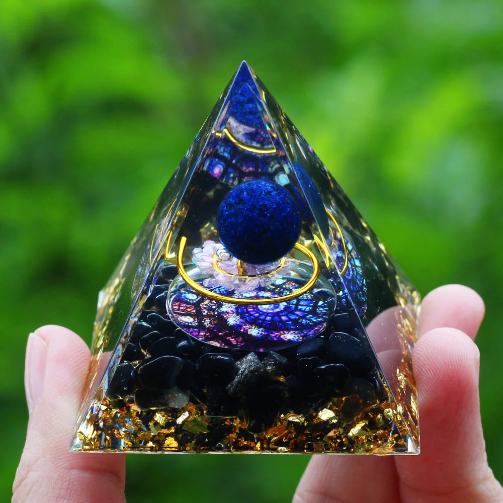Pyramid-shaped crystal with blue sphere and gold accents held by a hand against a green blurred background