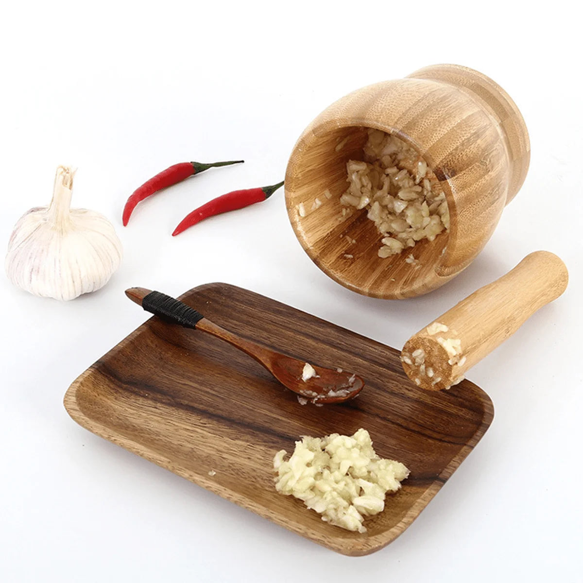 Wooden mortar and pestle with ground garlic on a wooden tray, surrounded by garlic and red peppers.