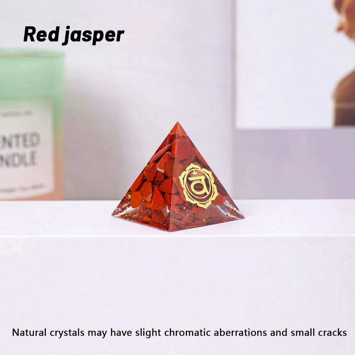 Red jasper pyramid crystal with a gold emblem on a white surface, with a candle and box in the background.