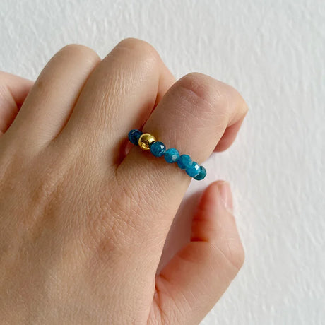 Hand wearing a ring with blue beads and gold accent on a white background
